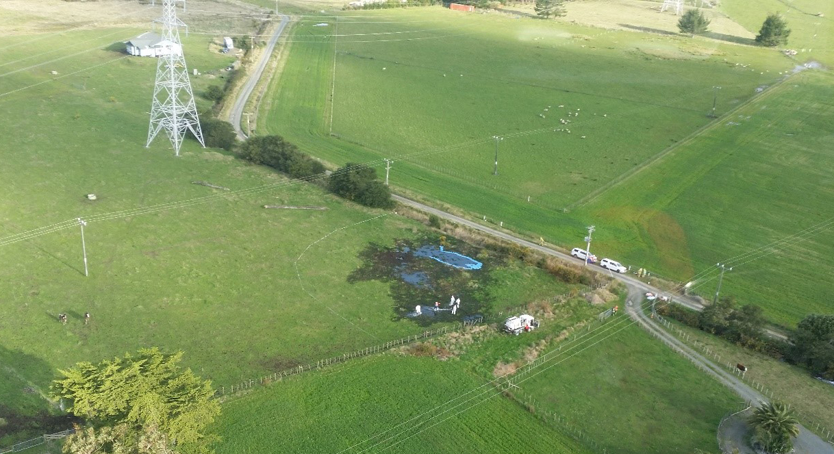 Aerial photographs showing investigation work at the site of the leak. This is the same field from Figure 4 and in the same area which appeared to have had earthworks completed. The pylon, road, and house are all visible as landmarks. There is a large area where the fuel has soaked the ground causing it to turn a black colour. In some areas a neon blue liquid has formed pools, this appears to be areas with a higher concentration of jet fuel.