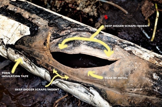 A photograph showing the damaged section of the pipeline in situ following excavation. This damage mainly consists of a large crescent shaped tear in the metal pipe. Also highlighted on the picture are 2 deep digger scrapes/indents and some torn insulation tape.